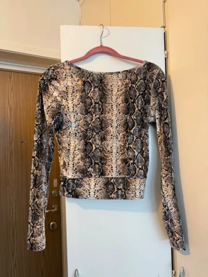 Gina tricot topp ormskinnsmönster - Långärmad topp med ormskinnsmönster i svart, beige och vitt. Toppen är i sammetsmaterial. Toppen har en rundad rygg och en figurnära passform.