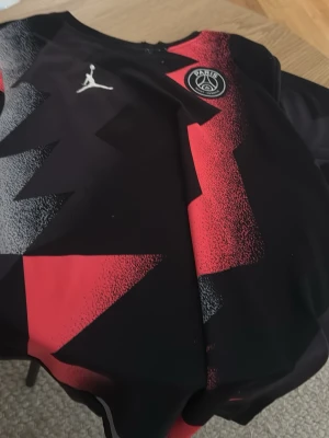 PSG x Jordan svart/röd träningströja - Snygg svart träningströja från PSG x Jordan med grafiskt rött och grått mönster. Tröjan har rund hals, tryckt Jumpman-logga och PSG-emblem på bröstet. Perfekt för fotbollsträning eller streetwear.