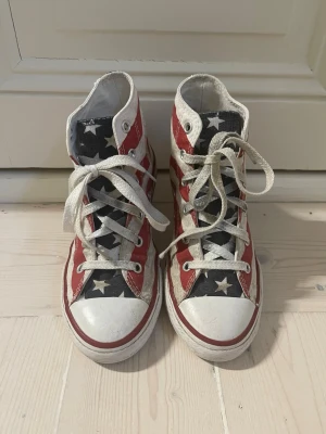 Converse USA-mönstrade high tops - Säljer ett par Converse high tops med amerikanskt flaggmönster i rött, vitt och blått med vita stjärnor. Klassisk canvasmodell med vita gummisulor och tåhätta. Skorna har vita snören och metallöljetter. Perfekta för dig som vill sticka ut med en unik sneaker.