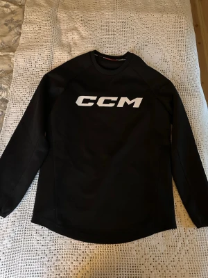 Svart långärmad tröja från CCM - Svart långärmad sporttröja från CCM med vit logga på bröstet och mindre logga i nacken. Tröjan har rund hals och är tillverkad i ett stretchigt funktionsmaterial, perfekt för träning eller hockey.