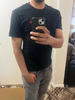 Svart t-shirt från Polo Ralph Lauren - Snygg svart t-shirt från Polo Ralph Lauren med klassisk röd logga broderad på bröstet. T-shirten har rund halsringning och korta ärmar. Tillverkad i mjuk bomull som känns skön mot huden. Perfekt till jeans eller shorts för en clean look.