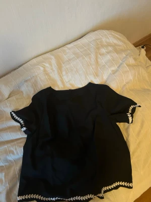 Svart blus med vit brodyrkant - Stilren svart blus med korta ärmar och rund halsringning. Blusen har dekorativa vita brodyrdetaljer längs ärmslut och nederkant som ger en trendig touch. Perfekt att matcha med både jeans och kjol för en snygg look.