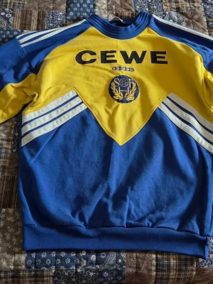 Vm 94 - Retroinspirerad blå och gul hoodie från Adidas med CEWE-tryck och SÖF-emblem på bröstet. Klassiska vita ränder på ärmarna och texten 'SVERIGE' i gult på ryggen. Tillverkad i mjukt bomullsmaterial och har ribbade muddar vid ärmslut och nederkant.