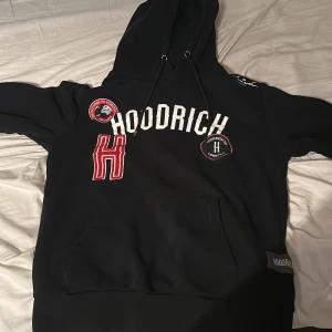 Svart hoodie från Hoodrich med tryck - Svart hoodie från Hoodrich med stor vit logga och röda detaljer framtill. Tröjan har luva med snöre och en stor magficka. Flera broderade patches och tryck ger en streetwear vibe. Perfekt för dig som gillar en cool och avslappnad stil.