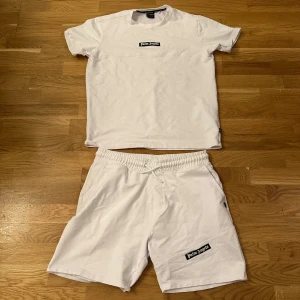 Vit t-shirt och shorts från Palm Angels - Snygg vit t-shirt från Palm Angels med svart logotyp på bröstet. Klassisk rund hals och korta ärmar, perfekt för en clean och fräsch look. Materialet känns mjukt och skönt mot huden, passar dig som gillar streetwear och stilrena plagg.