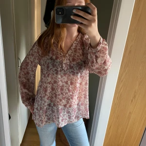 Blommig blus i ljusrosa och vinrött - Säljer en luftig blus med blommigt vinrött mönster på ljusrosa botten. Blusen har v-ringning, långa ärmar med resår vid handlederna och är gjord i ett tunt, flowy material. Perfekt för dig som gillar romantiska vibes och snygga detaljer.