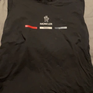 Svart Moncler Paris t-shirt - Svart t-shirt från Moncler med tryck framtill i vitt, rött och blått samt texten 'Moncler Paris'. Klassisk rund halsringning och korta ärmar. Skön och stilren modell i bomull, perfekt för dig som gillar streetwear med exklusiv känsla.