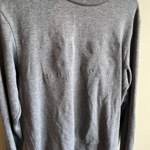 Grå slim fit tröja från Hugo Boss - Snygg grå tröja från Hugo Boss med diskret broderad logga på bröstet. Slim fit-modell med rund hals och långa ärmar. Mjuk och skön bomullskvalitet som passar perfekt till jeans eller chinos. Stilren och enkel design för en clean look.