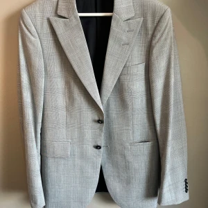 Tiger of Sweden kavaj  - Snygg grå kavaj från Tiger of Sweden med diskret rutigt mönster och klassisk passform. Kavajen har två knappar framtill, spetsiga slag och flera fickor både utvändigt och invändigt. Fodret är svart och materialet känns lyxigt och lätt. Perfekt för dig som gillar stilrena detaljer.