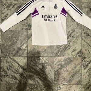 Vit långärmad Real Madrid fotbollströja - Säljer en vit långärmad Real Madrid fotbollströja från Adidas med svarta och lila detaljer. Tröjan har dragkedja vid halsen, svarta ränder på axlarna och klubbmärke på bröstet. Materialet är lätt och andas, perfekt för träning eller match.