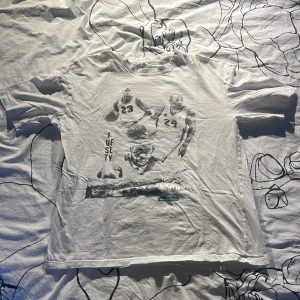 Vit t-shirt med baskettryck Jack & Jones - Vit t-shirt från Jack & Jones med coolt basketmotiv på både fram- och baksidan, featuring legendariska spelare. Rund hals och korta ärmar. Materialet är mjuk bomull, perfekt för dig som gillar sportiga och grafiska prints. Det finns en pytteliten fläck längst ner åt vänster på framsidan. Priset kan diskuteras. 