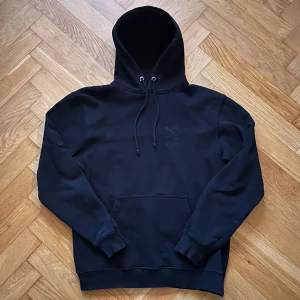 Svart St. Pauli hoodie med döskalle - Svart hoodie från St. Pauli i storlek S med klassisk döskalle- och korslagda ben-motiv på ryggen. Tröjan har huva med snörning, stor magficka och ribbade muddar. Materialet är mjuk bomull, perfekt för chill dagar. Snygg och enkel design med cool attityd.