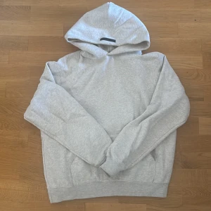 Grå Essentials Fear of God hoodie - Säljer en grå hoodie från Essentials Fear of God med svart logga på bröstet och en liten svart patch på huvan. Tröjan har lång ärm, huva och en klassisk loose passform. Perfekt för chill dagar och streetwear-stil.