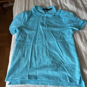 Turkos Polo Ralph Lauren t-shirt - Turkos slim fit t-shirt från Polo Ralph Lauren med klassisk pikékrage och korta ärmar. Materialet är bomull och på bröstet finns den ikoniska rosa broderade loggan. Två knappar vid halsen och diskreta slitsar i sidorna. Perfekt för en chill och stilren look.