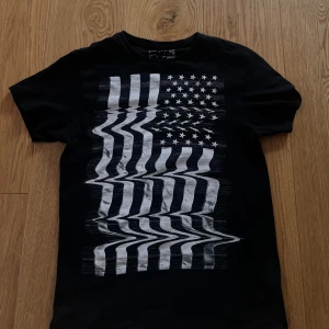 Svart t-shirt med glitchad flagga - Svart t-shirt med ett stort tryck av en glitchad amerikansk flagga i vitt på framsidan. T-shirten har rund hals och korta ärmar. Sitter slim. Fet tisha ska ej ljuga🔥