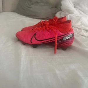 Nike rosa fotbollsskor med snörning - Säljer ett par grymt snygga fotbollsskor från Nike i stark rosa färg med svarta detaljer och logga på sidan. Skorna har snörning och en strumpliknande form runt ankeln för extra support. Ovandelen är i syntetmaterial och sulan har dobbar för bra grepp på planen.