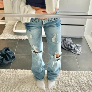 Ljusblå slitna bootcut jeans - Säljer ett par ljusblå jeans med bootcut passform och snygga slitningar över knäna och låren. Jeansen har låg midja, klassiska fickor med guldiga knappar bak och är tillverkade i jeansmaterial. Perfekta för en avslappnad och trendig look.