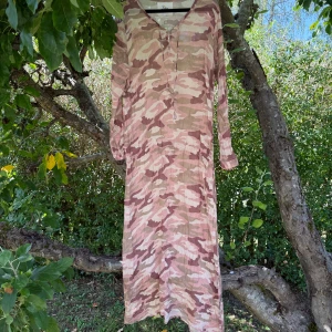 Lång camo-klänning från Fira - Säljer en långärmad klänning från Fira i storlek L med ett snyggt rosa och beige camouflage-mönster. Klänningen har v-ringning med snörning framtill och är gjord i ett lätt, luftigt material som passar perfekt för varma dagar. Fint skick, mekan ha några små tecken på användning. 