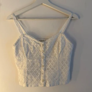 Hollister topp - Vit, ärmlös crop top med justerbara axelband och dekorativa knappar framtill. Toppen har en smickrande passform och är perfekt för varma dagar. Storlek S