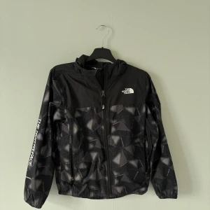 Svart vindjacka från The North Face - Snygg svart vindjacka från The North Face med geometriskt mönster i grått och vitt. Jackan har huva, dragkedja framtill och två fickor. Använd ett fåtal gånger så mycket bra skick! Säljer den för har helt enkelt vuxit ur den.