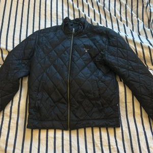 Svart quiltad jacka från GANT - Quiltad svart jacka från GANT i storlek S. Yttermaterialet är syntet och jackan har dragkedja framtill. Insidan har ett snyggt röd- och svart rutigt foder. Perfekt för dig som gillar klassisk stil med en modern twist. Perfekt nu till hösten 