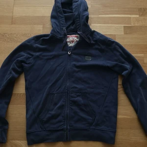 Marinblå hoodie från True Religion - Säljer en marinblå hoodie från True Religion med dragkedja och huva med snörning. Tröjan har två stora fickor framtill och en liten patch med logga på bröstet. Mått: underarmar=50cm längd runt 63cm