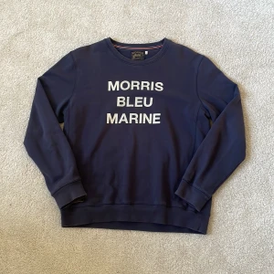Marinblå sweatshirt från Morris | L - Snygg marinblå sweatshirt från Morris i storlek L med vit text 'MORRIS BLEU MARINE' på bröstet. Tröjan har rund hals, ribbade muddar och är tillverkad i mjuk bomull. 