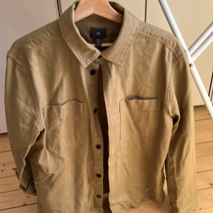 Beige overshirt från H&M i bomull - Snygg beige overshirt från H&M i storlek M. Skjortan har två stora bröstfickor, klassisk krage och knäppning med svarta knappar framtill. Tillverkad i slitstark bomull, perfekt att bära som lager över t-shirt. Enkel och stilren design.
