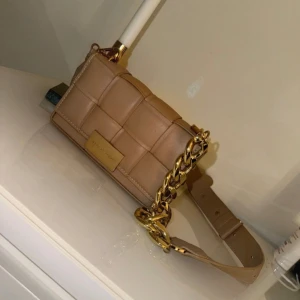 Beige quiltad axelväska från Steve Madden - Snygg beige axelväska från Steve Madden med quiltad design och bred axelrem. Väskan har en chunky guldfärgad kedjedetalj och en rektangulär form. Tillverkad i skinnimitation med justerbar rem och logoplatta framtill.