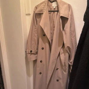 Beige trenchcoat - Snygg beige trenchcoat från H&M med klassisk krage, dubbelknäppning och justerbara remmar vid ärmsluten. Jackan har en loose passform och är tillverkad i bomullsmaterial. Perfekt för dig som gillar stilrena och tidlösa plagg. Storlek S men passar även M. Pris kan diskuteras 