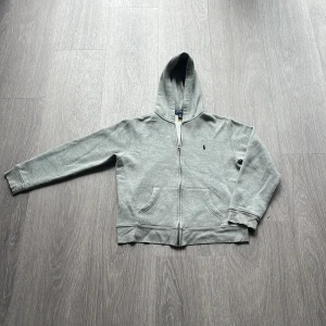 Grå zip hoodie från Polo Ralph Lauren - Grå hoodie från Polo Ralph Lauren med dragkedja och huva. Den är fet och perfekt för en vanlig outfit. Hör av er vid funderingar. Storleken är l 14-16 men skulle säga samma sak som cirka 164
