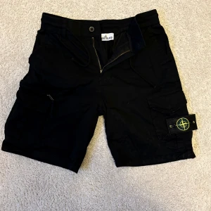 Svarta cargo shorts från Stone Island - Svarta cargo shorts från Stone Island med ikonisk patch på benet. De har flera fickor, dragkedja och knapp framtill samt snörning i midjan. Materialet är 97% bomull och 3% spandex för skön stretch. Perfekta för dig som gillar streetwear och funktionella detaljer.