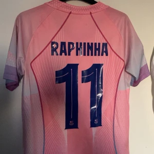 Barcelona Raphinha rosa matchtröja - Säljer en Barcelona fotbollströja med Raphinha och nummer 11 på ryggen. Tröjan är rosa med ljuslila detaljer, blått V-ringad krage och tryck från UEFA Foundation och Champions League på ärmen. Materialet är lätt och andas, Dri-Fit ADV. Spotify-logga på bröstet. Tröjan är knappt använd och finns inga tecken på användning eller defekter. Bara höra av sig vid frågor😌(Priset är inte hugget i sten) 
