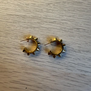 Guldiga Edblad örhängen med nitar - Säljer ett par coola guldiga örhängen från Edblad med chunky design och spetsiga nitar runt hela ringen. De har en halvcirkelform och ger en edgy vibe med sina metalliska detaljer. Perfekta för dig som gillar att sticka ut med accessoarer.