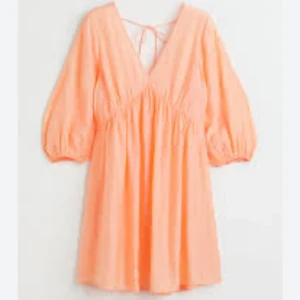 Persikofärgad kort klänning med v-ringning - Säljer en superfin persikofärgad kort klänning med djup v-ringning och puffiga långärmar. Klänningen har knyt i ryggen och är gjord i ett lätt, skrynkligt tyg som ger en chill och luftig känsla. Perfekt för varma dagar och snygg att styla med smycken.