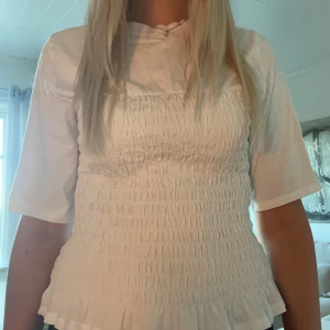 Smockad topp - Supersöt vit blus med smockad front och volangkant nedtill. Blusen har en hög hals och halvlånga ärmar som ger en chill vibe. Materialet känns som mjuk bomull och passformen är normal. Perfekt för dig som gillar stilrena och feminina detaljer.