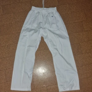 Vit karatekostym från Mizuno - Karatedräkt från Mizuno i helvitt med klassisk omlottjacka, byxa med snörning och tillhörande bälte. Tillverkad i slitstark bomull, perfekt för träning. Diskret Mizuno-logga på både jacka och byxa. Snygg och enkel design som passar dig som vill ha en clean look på mattan.