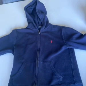 Marinblå hoodie från Polo Ralph Lauren - Snygg marinblå hoodie från Polo Ralph Lauren med dragkedja och huva. Tröjan har det klassiska röda Polo-logot broderat på bröstet och två praktiska fickor framtill. Perfekt för chill dagar och enkel att matcha med jeans eller joggers. Pris går att diskutera 