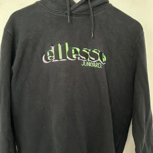 Svart hoodie från Ellesse Junkyard M - Svart hoodie från Ellesse Junkyard i storlek M. Tröjan har en stor logga i grönt och lila på bröstet, huva med snörning och långa ärmar. Mjuk insida och klassisk passform, perfekt för chill dagar. Snygg streetstyle-vibe med coolt tryck.