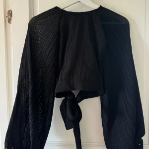 Svart omlott blus med knyt - Säljer en svart omlott blus med långa, transparenta ärmar och snyggt plisserat tyg. Blusen har ett knyt framtill och en öppen rygg, vilket ger en cool och trendig vibe. Perfekt för dig som gillar statement-plagg med lite extra edge.