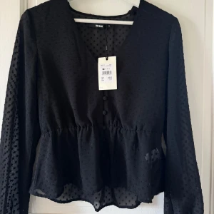 Svart prickig blus från Bik Bok - Snygg svart blus från Bik Bok med prickigt mönster och v-ringning. Blusen har långa ärmar, knappar framtill och ett lätt transparent material som ger en cool vibe. Midjan är lätt rynkad för en feminin siluett. Perfekt att styla med jeans eller kjol.