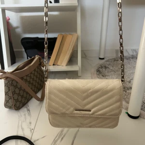 Vit quiltad axelväska från Bershka - Snygg vit axelväska från Bershka med quiltad design och kedjerem i metall. Väskan har ett stilrent, rakt lock med magnetknäppning och en insida med dragkedjefack. Perfekt storlek för mobilen och det viktigaste. Fräsch och trendig look.