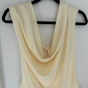 Ljusgul topp med snörning och öppen rygg - Säljer en champagnefärgad kortklänning med snygg omlott-design och draperad rygg. Klänningen har snörning i ryggen och är ärmlös, vilket ger en cool och modern vibe. Perfekt för dig som gillar ljusa färger och stilrena detaljer.