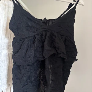 Svart volangig linnetopp med smala band - HELT NY, UTAN PRISLAPP! Svart linnetopp med smala axelband och snygga volangdetaljer framtill. Toppen har en skrynklig textur och v-ringad hals, vilket ger en cool och avslappnad vibe. Perfekt för dig som gillar att sticka ut med lager på lager och mjuka material.