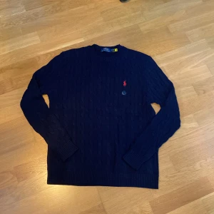 Mörkblå stickad tröja från Polo Ralph Lauren - Säljer en snygg mörkblå stickad tröja från Polo Ralph Lauren med det klassiska röda broderade logotypen på bröstet. Tröjan har en kabelstickad design och långa ärmar. Köpte denna vara på Vinted. Nypris runt 2000kr.