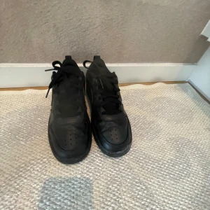 Svarta Nike sneakers storlek 35  - Säljer ett par helsvarta Nike sneakers med klassisk låg siluett och rund tå. Skorna har snygg snörning, platt sula och är tillverkade i skinn med diskret Nike-logga på sidan. Perfekta för dig som gillar stilrena och tidlösa sneakers.