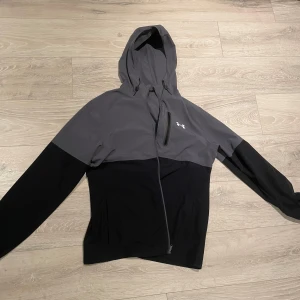 Under Armour grå och svart vindjacka - Snygg vindjacka från Under Armour i grått och svart med huva och snörning. Jackan har en cool asymmetrisk dragkedja och en bröstficka med zip. Perfekt för dig som gillar sportig stil och vill ha något funktionellt och stilrent. BYXORNA FINNS OCKSÅ HELA TRACKSUITEN 1200KR!