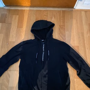 Svart hoodie från Armani Exchange XS - Svart hoodie från Armani Exchange i storlek XS. Tröjan har huva med snörning där loggan syns, långa ärmar och fickor framtill. Materialet är mjuk bomull och polyester, perfekt för chill dagar. Klassisk och stilren look med ribbad nederkant.