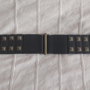 Svart midjebälte/skärp med nitar och resår - Brett svart midjebälte med coola fyrkantiga guldfärgade nitar och metallspänne. Bältet har en mix av elastiskt tyg och skinnimitation framtill, vilket ger både stil och komfort. Perfekt för att ge din outfit en edgy touch.