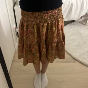 Mönstrad volangkjol i rosa och grönt - Supersöt kortkjol med volanger och smock i midjan. Kjolen har ett paisleyinspirerat mönster i rosa, grönt och inslag av beige. Lätt och flowy känsla, perfekt för varma dagar. Passar dig som gillar boho-vibe och vill ha något färgglatt och bekvämt.Storleken är 32 men kan göra större för att det är ett väldigt stretchat material 